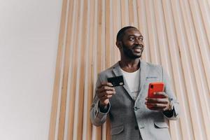 empresário afro-americano animado usando smartphone e cartão de crédito para fazer compras online foto