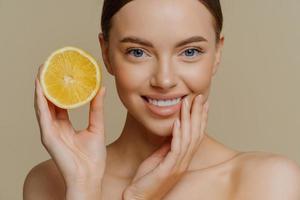 close-up tiro de mulher bonita e delicada sorri suavemente tem uma pele saudável e bem cuidada segura uma fatia de limão usa frutas cítricas para beleza e saúde fica sem camisa isolada sobre fundo marrom foto