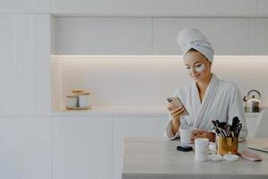 feliz jovem europeia vestida de roupão digita mensagens de texto no bate-papo on-line durante o intervalo para o café passa por procedimentos de cuidados com a pele aplica manchas de beleza poses sobre o interior da cozinha em casa foto