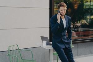 comerciante masculino bonito bem sucedido discutindo os preços das ações enquanto se inclina na mesa do café na calçada foto