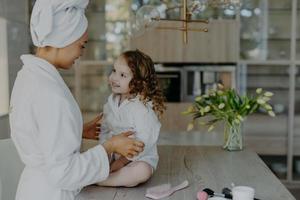 mãe e filha em roupões de banho brancos têm uma conversa agradável uma com a outra, passando por procedimentos de beleza depois de tomar banho. pequena menina adorável com cabelo encaracolado olha para a mamãe em casa. foto