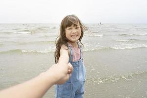 menina feliz puxa a mão dos pais para o mar com um grande sorriso e ondas no fundo, conceito de estilo de vida familiar foto