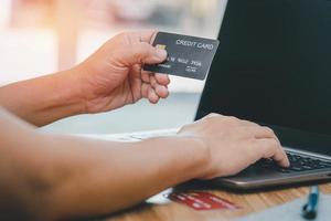 mão um homem segure o cartão de crédito digite o número do cartão no computador portátil na mesa de madeira para compras on-line pedido de compras em casa e conceito de entrega foto