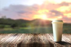 café quente em um copo de plástico para viagem em uma mesa de madeira marrom com vista para a natureza rural e montanha com fundo de céu agradável e nascer do sol de manhã, bebidas refrescantes e relaxantes nas férias. foto