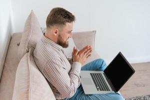 jovem atraente sentado no sofá em casa, trabalhando no laptop online, usando foto