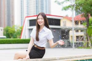 retrato de estudante tailandês adulto em uniforme de estudante universitário. menina bonita asiática sentado agitar a água e sorrindo alegremente na universidade ao ar livre.