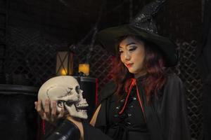 menina asiática em fantasia de bruxa segurando o crânio humano para o conceito de doces ou travessuras de festa de halloween com fundo preto escuro com espaço de cópia foto