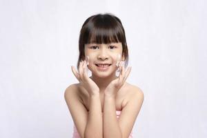 linda garota de ginástica asiática rindo aplicando creme para o rosto com a pele limpa e fresca no fundo branco. tratamentos faciais e spas, cuidados faciais, rosto de menina asiática foto