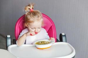 bebê infantil com cara suja comendo sopa com colher. menina bonitinha sentada na cadeira alta do bebê na cozinha em casa. conceito de infância de família de maternidade. foto