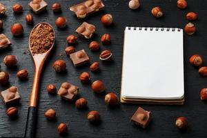 chocolate com avelãs, um caderno com páginas em branco e uma colher de pau com cacau em um fundo escuro, cercado por nozes com casca e descascadas. foto