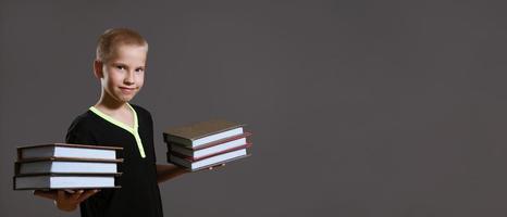 menino bonito segurando pilhas de livros em fundo cinza foto
