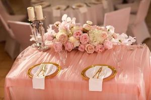 decoração de mesa com toalha de mesa rosa, castiçais de cristal com velas e flores rosa brancas no restaurante. dia de casamento elegante. mesa de ajuste com placa de ouro, garfo e faca foto