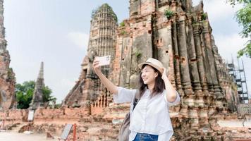 jovem viajante asiática usando telefone celular tomando selfie enquanto viaja sozinho no templo ayuthaya tailândia em dia ensolarado de verão. fêmea alegre desfruta de estilo de vida ao ar livre em férias foto