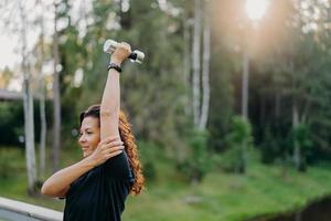 tiro horizontal de mulher morena motivada levanta o braço e segura o haltere, treina os músculos, tem treino regular de manhã, veste camiseta preta, posa ao ar livre contra o fundo das árvores. musculação foto