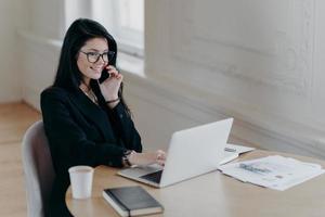 bela executiva feminina faz trabalho remoto, fala via telefone celular e verifica informações em poses de computador portátil no local de trabalho cercado por documentos de papel de café bloco de notas tem conversa telefônica foto