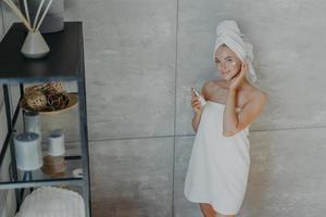 vista de cima da linda mulher encantadora fica perto da parede cinza no banheiro aplica loção facial embrulhada em toalha de banho branca goza de pele impecável usa produtos cosméticos cuidados com a pele foto