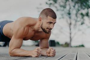 close-up tiro de homem motivado barbudo fica na prancha, treina músculos e quer ter um corpo forte. esportista fazendo exercícios ao ar livre. cara atlético faz flexões. conceito de esporte e estilo de vida foto