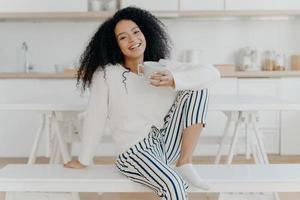 bela jovem fêmea com corte de cabelo afro, posa no banco branco interior, vestida com roupas elegantes, gosta de bebidas aromáticas, gosta de café da manhã em casa, posa contra o interior da cozinha turva foto