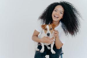 retrato de alegre garota encaracolada acariciando seu cachorro, regozijando-se comprando jack russell terrier, sorri amplamente, brinca com animais, usa roupas casuais, isoladas sobre fundo branco, goza de bom dia foto