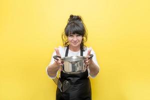 mulher bonita sorridente em um avental preto com um batedor e uma espátula segura uma panela, fundo amarelo, o conceito de oferecer comida deliciosa e cozinhar foto