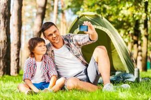 capturando os momentos brilhantes. alegre pai e filho sentados perto da barraca enquanto acampam na floresta foto