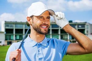 golfista confiante. vista de ângulo baixo do jovem golfista feliz segurando o motorista e ajustando seu boné em pé no campo de golfe foto