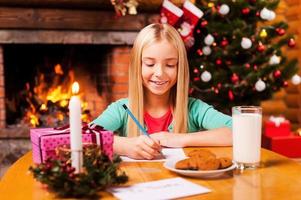 escrevendo uma carta para o papai noel. menina bonitinha escrevendo uma carta para o papai noel enquanto está sentado em casa com árvore de natal e lareira ao fundo foto