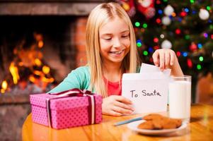 lista de desejos para o papai noel. menina bonitinha colocando uma carta para o papai noel no envelope enquanto está sentado em casa com árvore de natal e lareira ao fundo foto