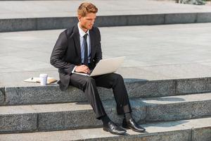 empresário trabalhando ao ar livre. jovem confiante de camisa e gravata trabalhando no laptop enquanto está sentado ao ar livre foto