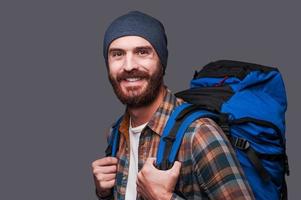 turista feliz. bonito jovem barbudo carregando mochila e sorrindo em pé contra um fundo cinza foto