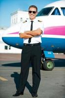 pronto para voar. comprimento total do piloto masculino confiante de uniforme, mantendo os braços cruzados e sorrindo com o avião ao fundo foto