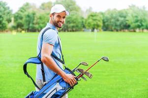 pronto para jogar. vista traseira do jovem golfista feliz carregando saco de golfe com motoristas e olhando por cima do ombro em pé no campo de golfe foto