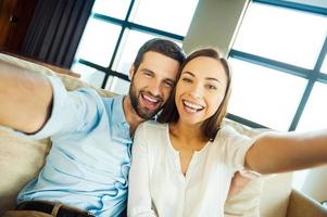 capturando diversão. lindo jovem casal amoroso se unindo e fazendo selfie enquanto está sentado no sofá juntos foto