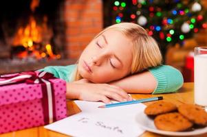 pequeno sonhador. menina bonitinha dormindo enquanto inclina a cabeça na mesa com árvore de natal e lareira ao fundo foto