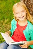 lindo leitor de livros. vista superior da menina de cabelo loiro bonitinho segurando o livro e sorrindo para você enquanto está sentado na grama verde e inclinando-se para a árvore foto