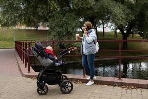 mãe adulta caminha com um bebê em um carrinho de bebê em um parque da cidade falando em um telefone celular foto