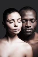 um amor. homem africano sem camisa e mulher caucasiana, unindo seus rostos um ao outro e mantendo os olhos fechados em pé contra um fundo cinza foto