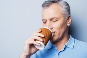 bebendo café fresco. homem sênior bonito segurando a xícara de café e mantendo os olhos fechados em pé contra um fundo cinza foto