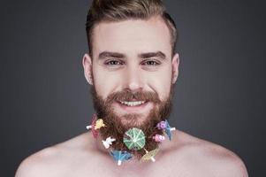 barba fofa. retrato de jovem bonito sem camisa com brinquedos e guarda-sóis de coquetel na barba olhando para a câmera e sorrindo em pé contra um fundo cinza foto