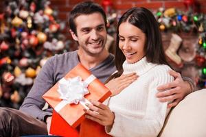 que grande surpresa bela jovem abrindo uma caixa de presente e sorrindo enquanto o namorado sentado perto dela no sofá com decoração de natal ao fundo