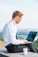 ter liberdade para trabalhar em qualquer lugar. vista traseira do jovem confiante de camisa e gravata trabalhando no laptop enquanto está sentado ao ar livre com a paisagem urbana ao fundo foto