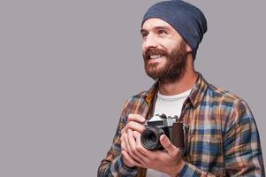 fotógrafo feliz. bonito jovem barbudo segurando a câmera antiquada e desviar o olhar com um sorriso em pé contra um fundo cinza foto