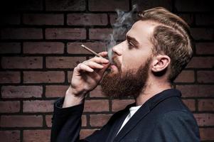 em seu próprio estilo. Vista lateral do jovem barbudo bonito fumando um cigarro em pé contra a parede de tijolos
