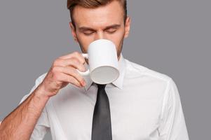 finalmente pausa para o café. jovem cansado em trajes formais segurando a xícara de café e bebendo em pé contra um fundo cinza foto