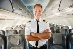 este é o meu avião. piloto masculino confiante de uniforme, mantendo os braços cruzados e sorrindo em pé dentro do avião foto