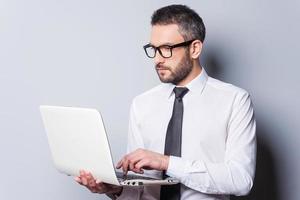 especialista em negócios no trabalho. homem maduro confiante de camisa e gravata trabalhando no laptop em pé contra um fundo cinza foto