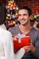 que surpresa vista traseira do jovem dando uma caixa de presente vermelha para sua namorada com árvore de natal ao fundo foto