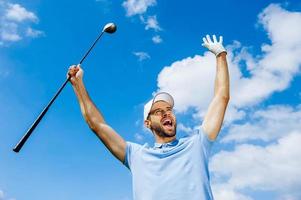 vista de ângulo baixo do jovem golfista feliz segurando o motorista e levantando os braços com céu azul como pano de fundo foto