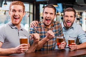 é um objetivo três jovens felizes em roupas casuais segurando copos com cerveja e torcendo enquanto assistem a uma partida de futebol no bar juntos