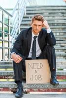 preciso de um emprego. jovem deprimido em trajes formais segurando cartaz com mensagem de texto de trabalho enquanto está sentado na escada foto